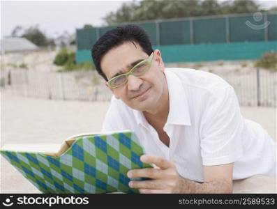 Portrait of a mature man lying on sand and holding a book