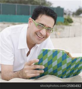 Portrait of a mature man lying on sand and holding a book