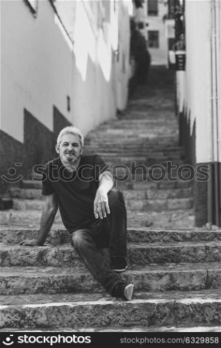 Portrait of a mature man sitting on steps in the street. Senior male with white hair and beard wearing casual clothes in urban background.