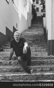 Portrait of a mature man sitting on steps in the street. Senior male with white hair and beard wearing casual clothes in urban background.