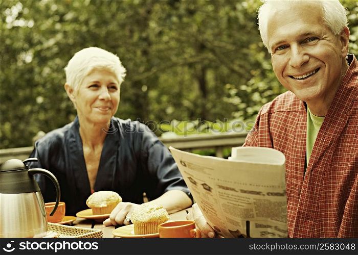 Portrait of a mature man smiling and holding a newspaper with a senior woman in the background