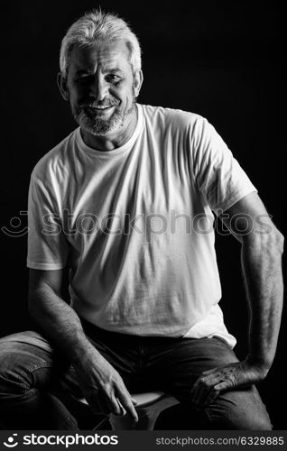 Portrait of a mature man smiling looking at camera. Senior male with white hair and beard laughing wearing casual clothes isolated on black background. Studio shot in black and white.