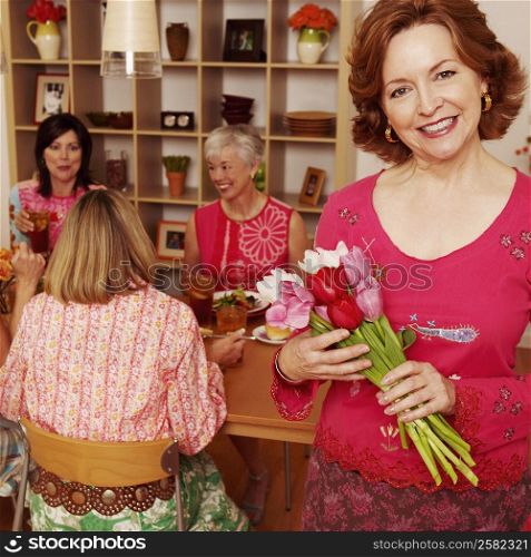 Portrait of a mature woman holding a bouquet of flowers and her friends sitting at the dining table in the background