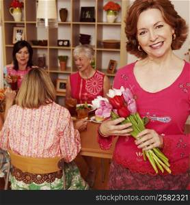 Portrait of a mature woman holding a bouquet of flowers and her friends sitting at the dining table in the background