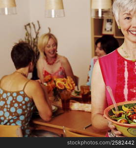 Portrait of a mature woman holding a bowl of salad and her friends sitting at the dining table in the background