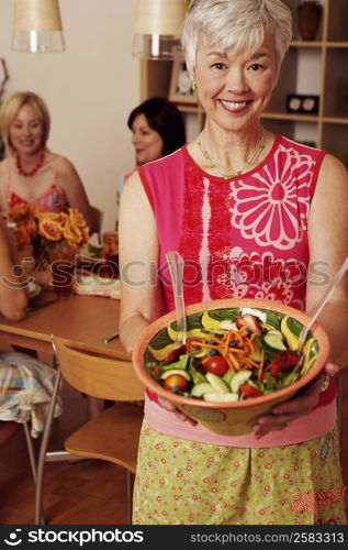 Portrait of a mature woman holding a bowl of salad and her friends sitting at the dining table in the background