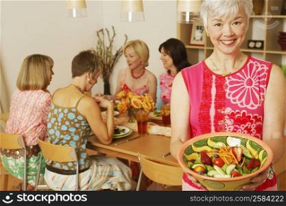 Portrait of a mature woman holding a bowl of salad and her friends sitting at the dining table in the background