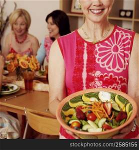 Portrait of a mature woman holding a bowl of salad and her friends sitting at the dining table in the background