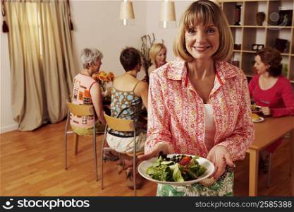 Portrait of a mature woman holding a plate of salad and her friends sitting behind her