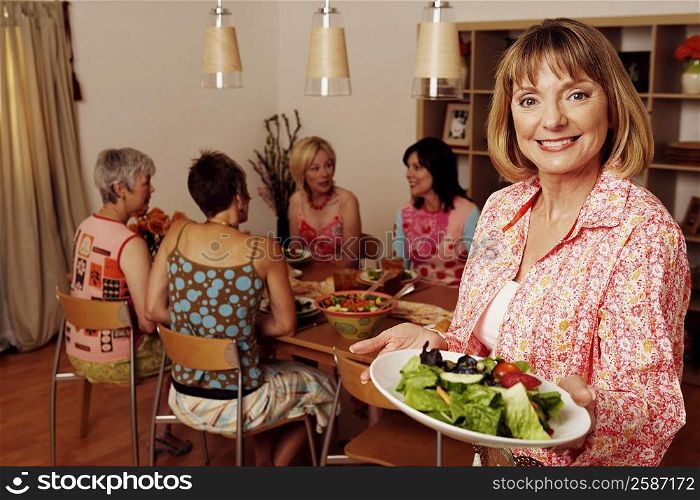 Portrait of a mature woman holding a plate of salad and her friends sitting at a dining table in the background