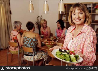 Portrait of a mature woman holding a plate of salad and her friends sitting at a dining table in the background