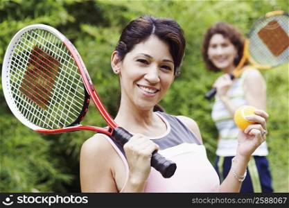 Portrait of a mature woman holding a tennis ball and a tennis racket with another mature woman standing behind her