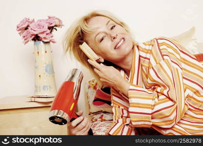 Portrait of a mature woman lying in bed and drying her hair with a hair dryer