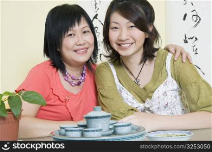 Portrait of a mature woman sitting with her arm around her daughter at a table and smiling