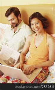 Portrait of a mature woman using a laptop with a mature man reading a newspaper beside her