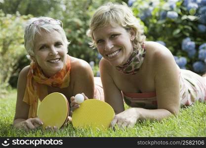 Portrait of a mature woman with her sister lying on grass and holding table tennis rackets