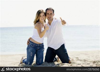 Portrait of a mid adult man and a young woman kneeling on the beach and showing Thumbs Up sign