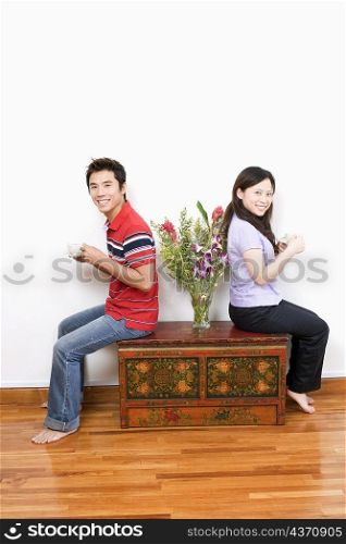 Portrait of a mid adult man and a young woman sitting on a trunk and holding cups of tea