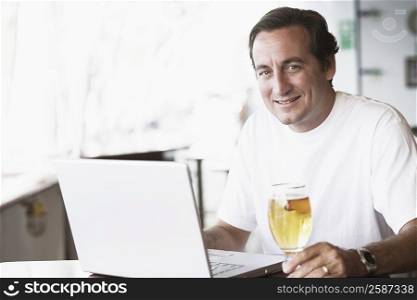 Portrait of a mid adult man sitting in front of a laptop and holding a glass of beer