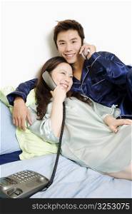 Portrait of a mid adult man using a mobile phone with a young woman talking on the telephone on the bed