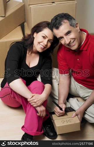Portrait of a mid adult man writing on a cardboard box with a mid adult woman sitting beside him