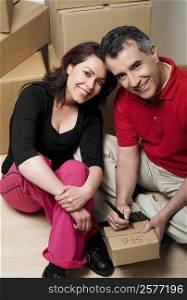 Portrait of a mid adult man writing on a cardboard box with a mid adult woman sitting beside him