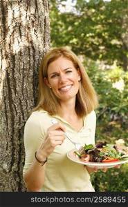 Portrait of a mid adult woman leaning against a tree and eating a vegetable salad