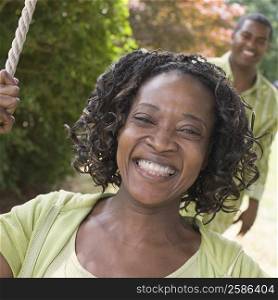 Portrait of a mid adult woman sitting on a rope swing and smiling with a mature man in the background