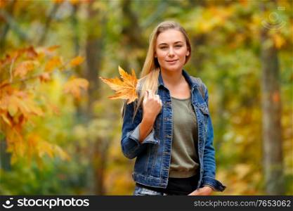 Portrait of a nice blond female holding in hand beautiful dry yellow maple leaf over autumnal foliage background, having fun in the autumn park
