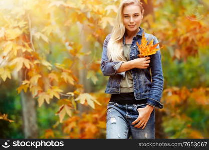 Portrait of a nice blond female holding in hand beautiful dry yellow maple leaf over autumnal foliage background, having fun in the autumn park in warm sunny day