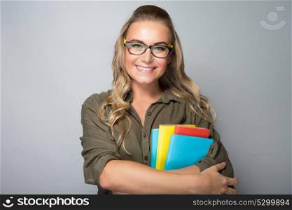 Portrait of a nice blond student girl wearing glasses, holding colorful books, shot over gray background, education in university