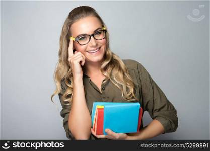 Portrait of a nice blond student girl with colorful books in hands wearing glasses, over gray background, education in university