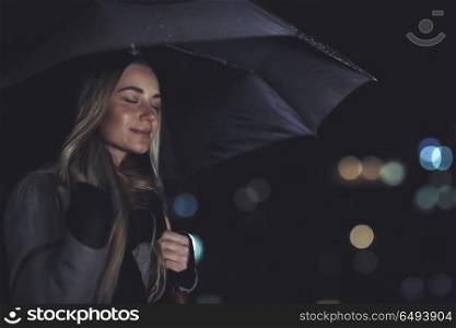 Portrait of a nice calm female with closed eyes and smile standing outdoors at night, enjoys the sound of the rain drumming on the umbrella, peacefulness concept. Calm female enjoying rainy night