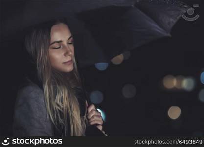 Portrait of a nice calm female with closed eyes standing outdoors at night, enjoys the sound of the rain drumming on the umbrella, peacefulness concept. Calm female under the rain at night