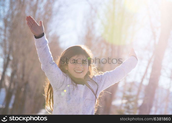 Portrait of a nice cheerful little girl running with raised up hands enjoying winter holidays in the park, photo with sun flare, happy carefree childhood