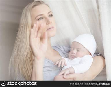 Portrait of a nice gentle mommy with sleeping newborn baby at home, looking through the window, usual domestic life
