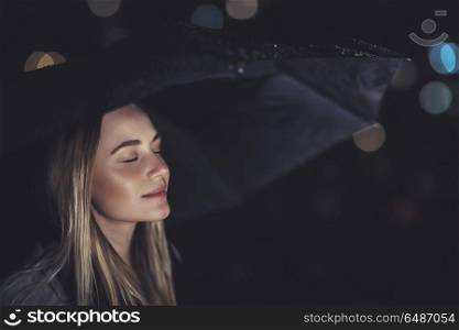 Portrait of a nice peaceful woman closing eyes of pleasure, standing under umbrella at night and enjoying rain, peace and mind calmness concept. Woman enjoying rainy night