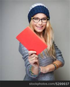 Portrait of a nice student girl with school book in hand over gray background, wearing stylish hat and glasses, education in the university