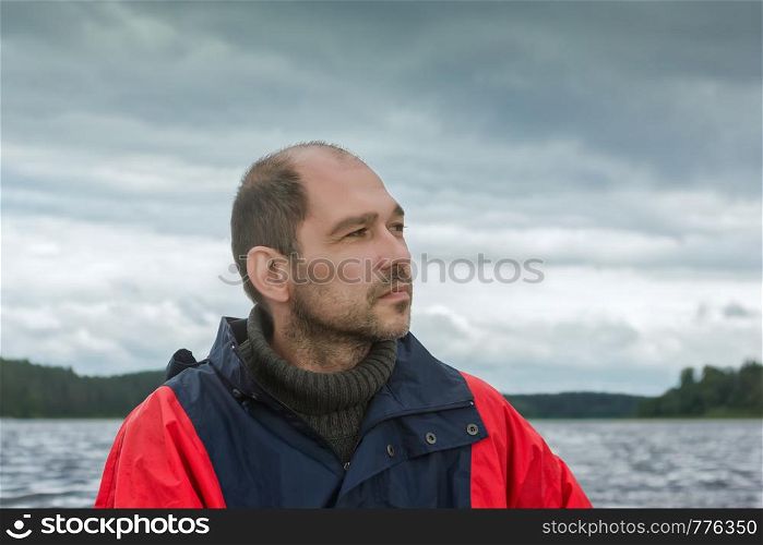 Portrait of a pensive bearded mature man against the overcast cloudy sky and the stormy lake. Selective focus on foreground, blurred natural background with copy space.