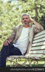 Portrait of a pensive mature man sitting on a bench in an urban park. Senior male with white hair and beard wearing casual clothes.