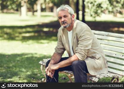 Portrait of a pensive mature man sitting on a bench in an urban park. Senior male with white hair and beard wearing casual clothes.