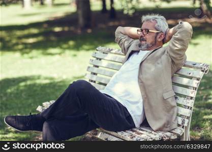 Portrait of a pensive mature man sitting on a bench in an urban park. Senior male with white hair and beard wearing casual clothes and eyesglasses.