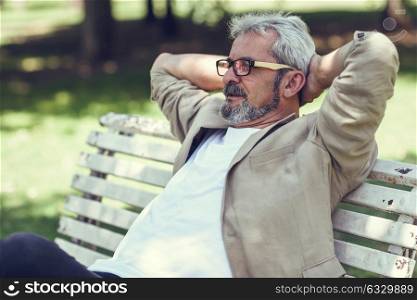 Portrait of a pensive mature man sitting on a bench in an urban park. Senior male with white hair and beard smiling wearing casual clothes. Old man wearing eyeglasses.