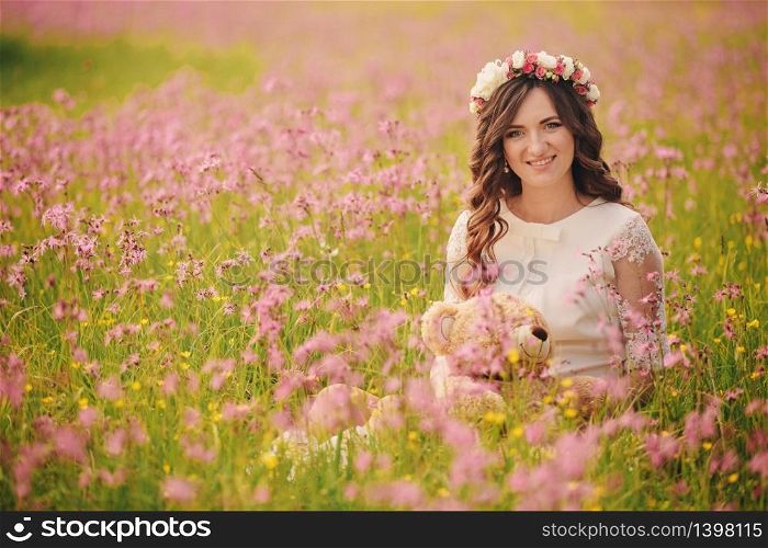 Portrait of a pregnant woman with teddy bear in fieid of pink flowers. Young beautiful pregnant girl with a wreath on her head in the sun. Motherhood. Spring. copy space. selective focus.. Portrait of a pregnant woman with teddy bear in fieid of pink flowers. Young beautiful pregnant girl with a wreath on her head in the sun. Motherhood. Spring. copy space. selective focus