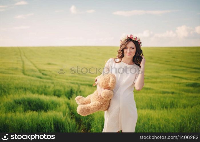 Portrait of a pregnant woman with teddy bear in hand in the field of grass. Young beautiful pregnant girl with a wreath on her head in the sun. Motherhood. Spring. copy space. selective focus.. Portrait of a pregnant woman with teddy bear in hand in the field of grass. Young beautiful pregnant girl with a wreath on her head in the sun. Motherhood. Spring. copy space. selective focus