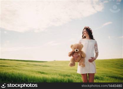 Portrait of a pregnant woman with teddy bear in hand in the field of grass. Young beautiful pregnant girl with a wreath on her head in the sun. Motherhood. Spring. copy space. selective focus.. Portrait of a pregnant woman with teddy bear in hand in the field of grass. Young beautiful pregnant girl with a wreath on her head in the sun. Motherhood. Spring. copy space. selective focus