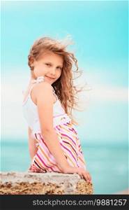 Portrait of a pretty little girl with waving in the wind long hair sitting on the beach against the blue sky