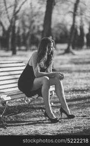Portrait of a pretty woman sitting in a bench in a urban park wearing black dress and high heels