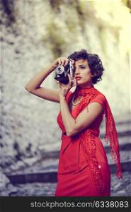 Portrait of a pretty woman, vintage style, in urban background, wearing a red dress taking photographs with a old camera