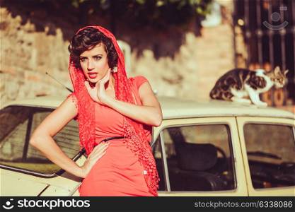 Portrait of a pretty woman, vintage style, in urban background, wearing a red dress, with a old car and a cat